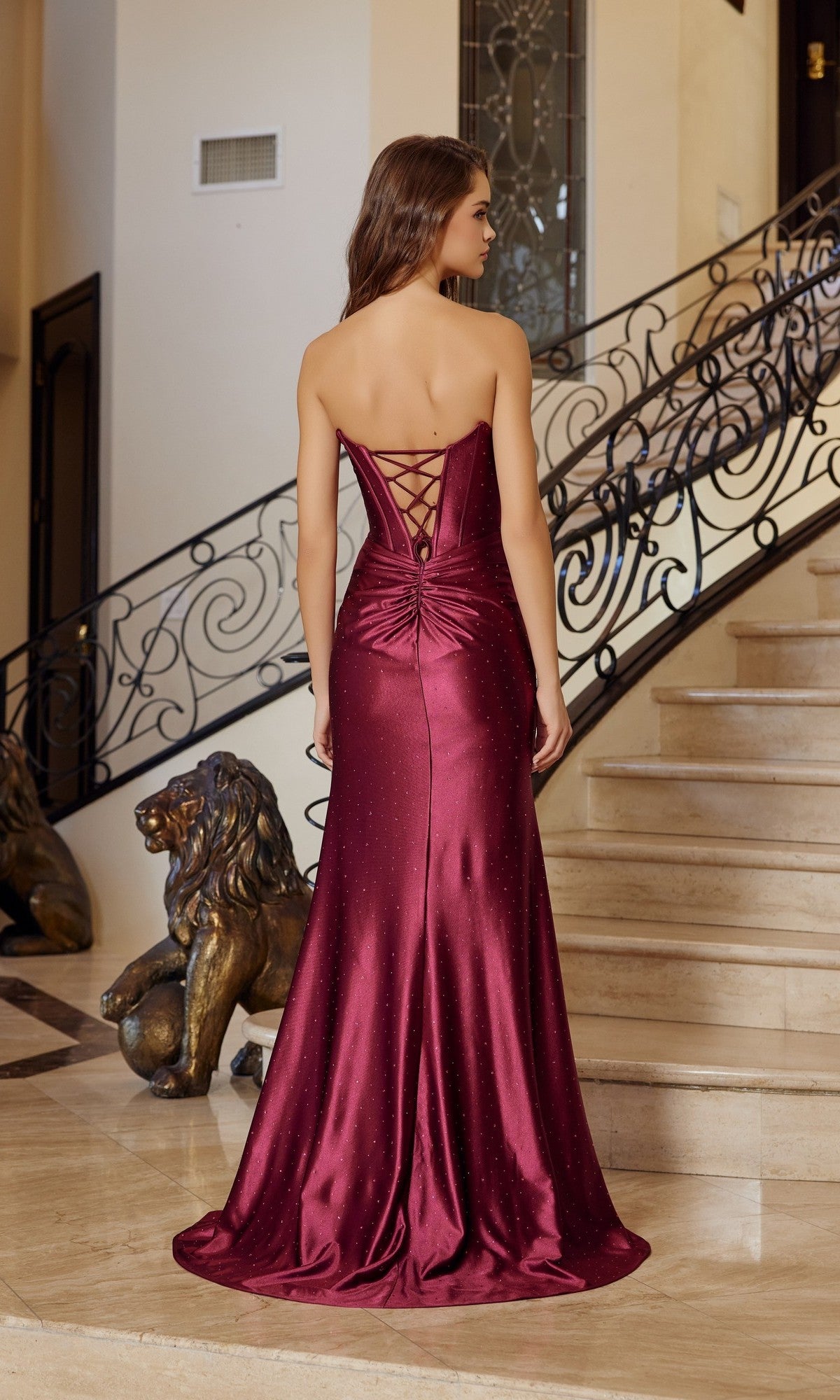 A woman in the Nox Anabel T1861 long prom dress—burgundy satin with rhinestone details and a lace-up corset bodice—stands at the base of a staircase in an elegant interior.